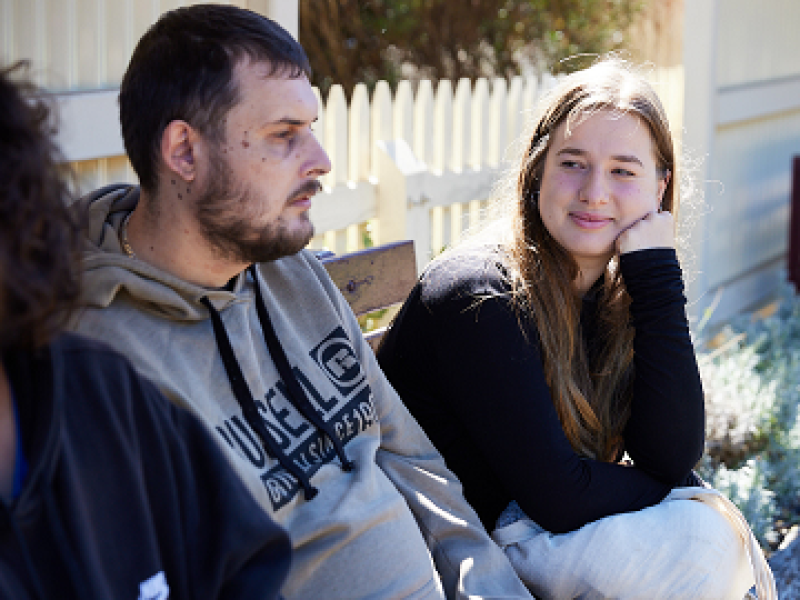 Man and woman sitting on a bench. Woman is resting her face on her hand. Man is looking forward.