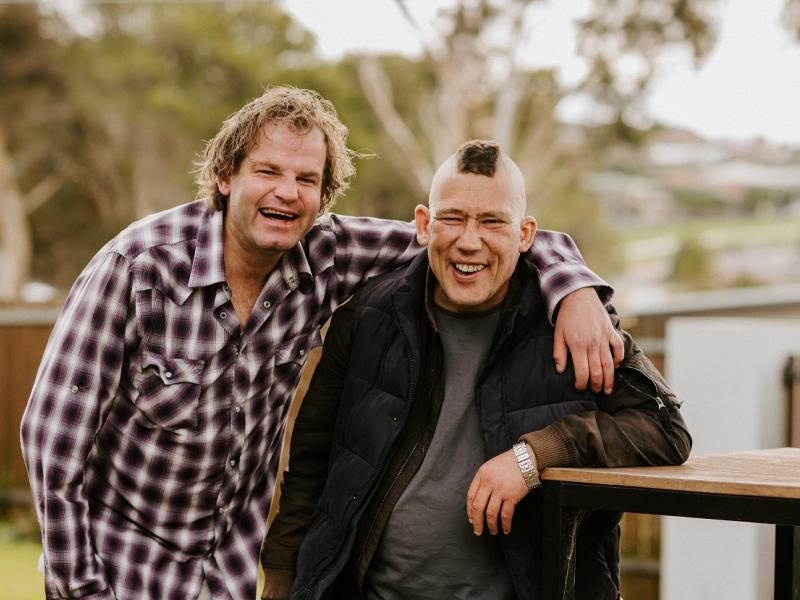 Two men smiling in a backyard. One man is embracing the other with his arm around the other.