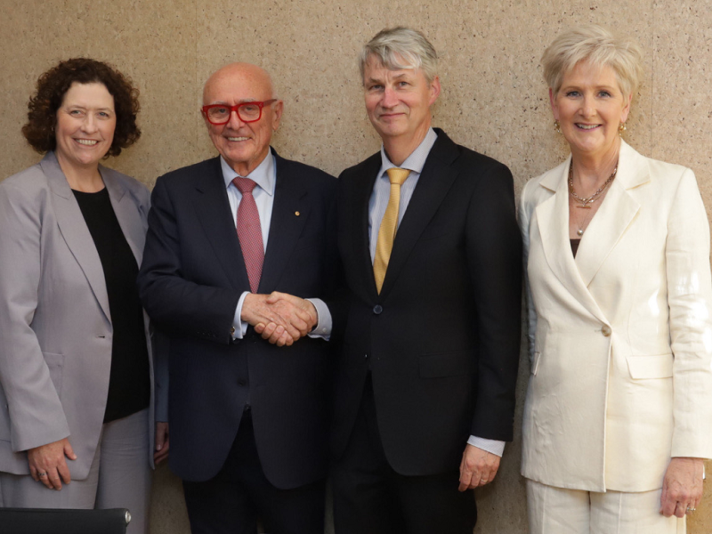 Four people standing and smiling at a wall. Two men are shaking hands.