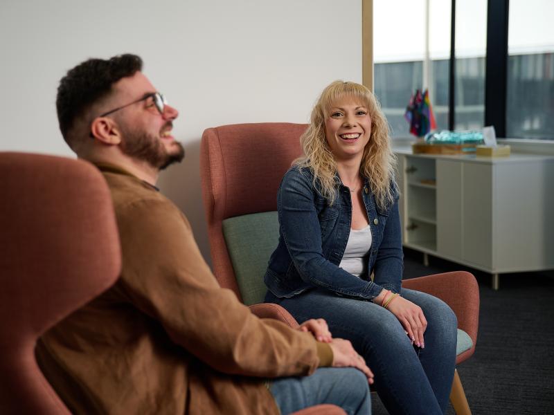 Two people sitting on lounge chairs having a chat. One person can be seen smiling and looking directly at the camera, while the other person can be seen smiling and laughing.