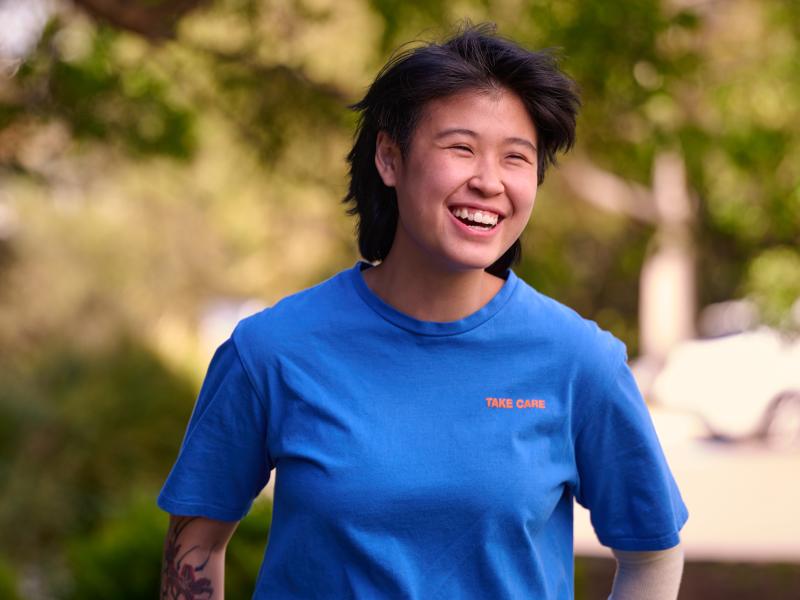 Smiling person in a blue T-shirt standing outdoors in a leafy garden.
