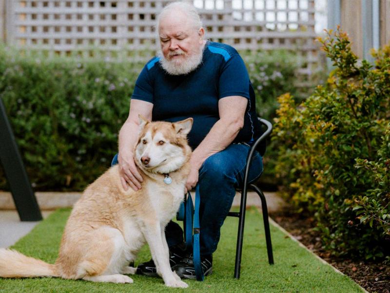 Person seated outdoors beside a large dog, with a hand resting on the dog’s back. 