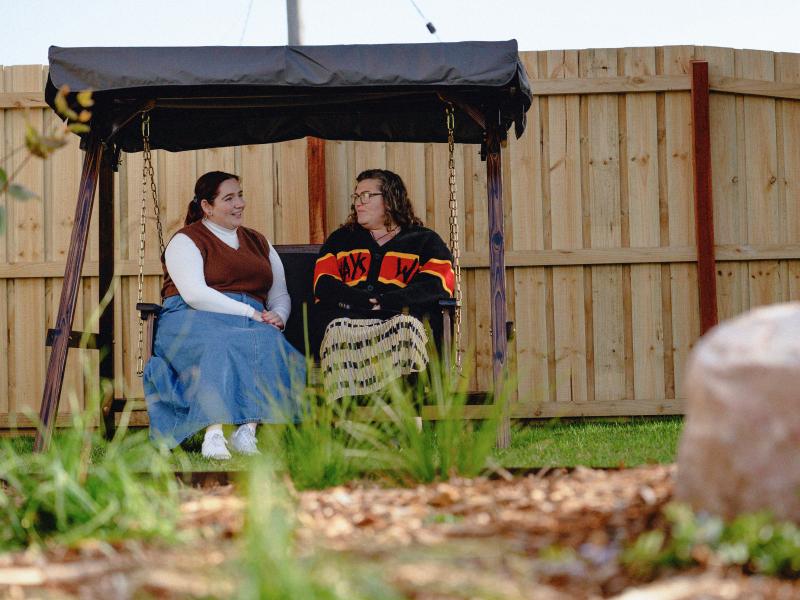 Two people sitting together on an outdoor swing seat, talking in a garden. 
