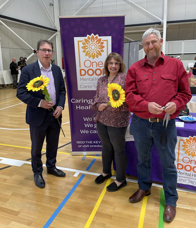 A man and woman holding gigantic sunflowers in front of a One Door banner